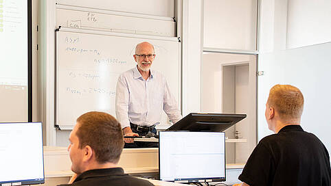 A trainer stands at a lectern in front of a whiteboard and explains