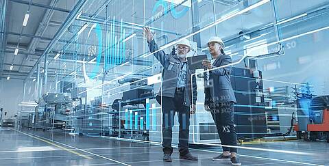 man and woman with safety helmets on in a production hall, looking at a virtual HMI dashboard