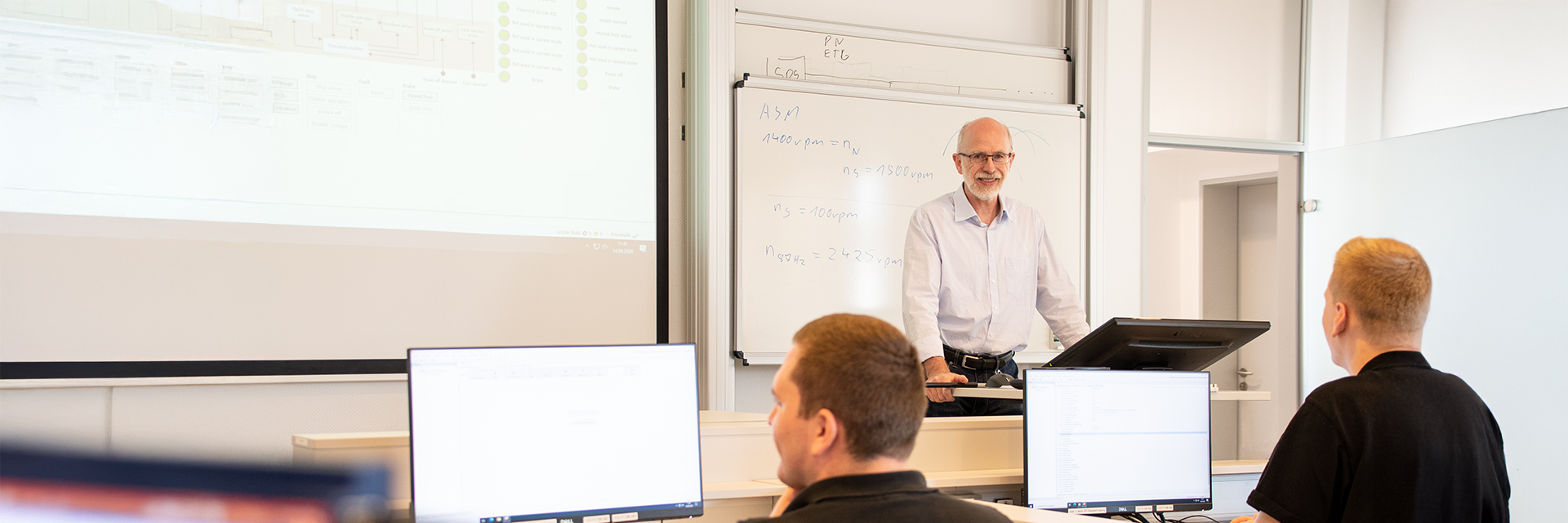 A trainer stands at a lectern in front of a whiteboard and explains